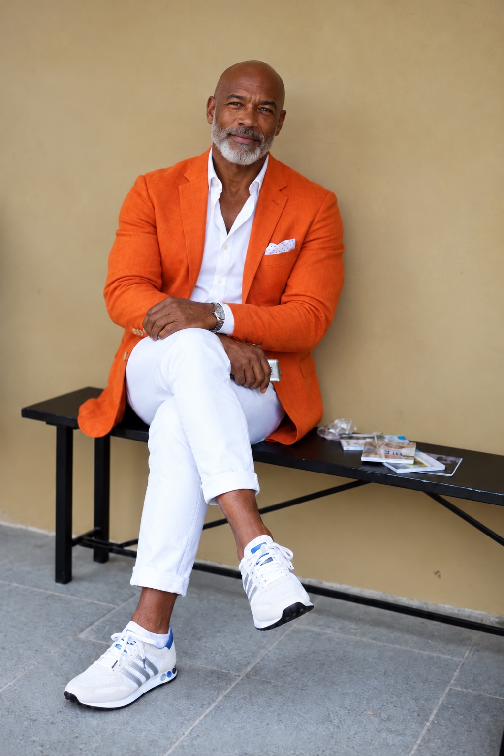 A bald Black man in his fifties with a grey beard, wearing an orange linen blazer, white shirt and white trousers, seated on a bench against a warm wall